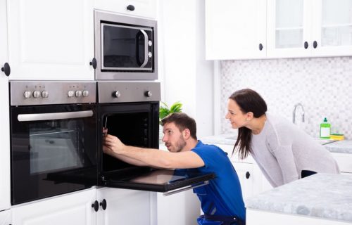 Smiling Young Woman Looking At Technician Fixing Oven In Kitchen